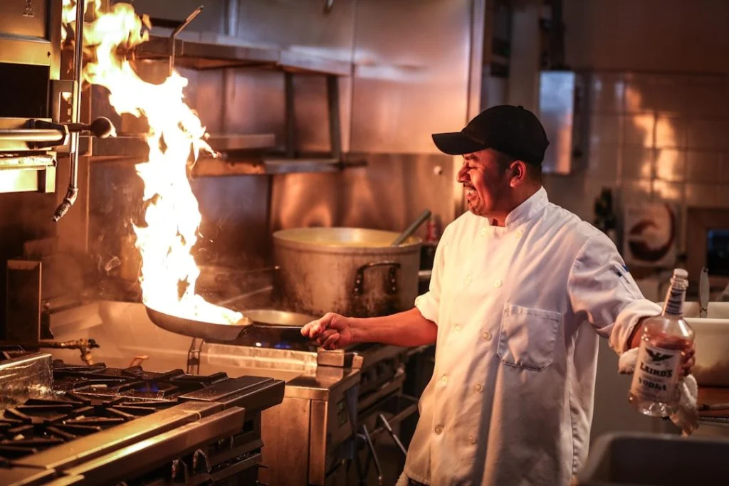 A chef in a white jacket controls a burst of flame on the line in a high-pressure restaurant kitchen, evoking the chaos and intensity of The Bear